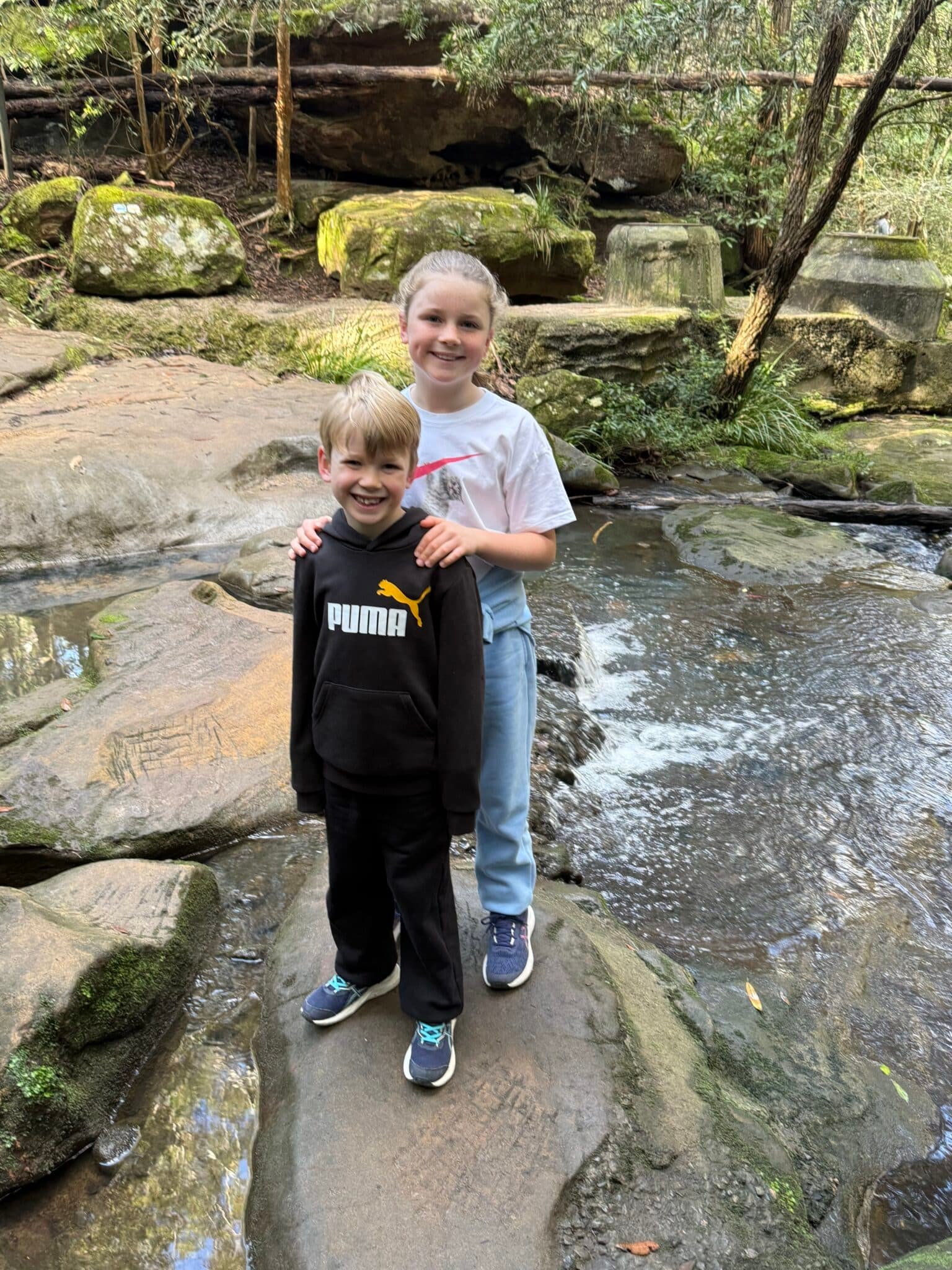 Smiling brother and sister standing on rocks by a creek in Sydney bushland, symbolising natural joy, family connection, and emotional wellbeing.