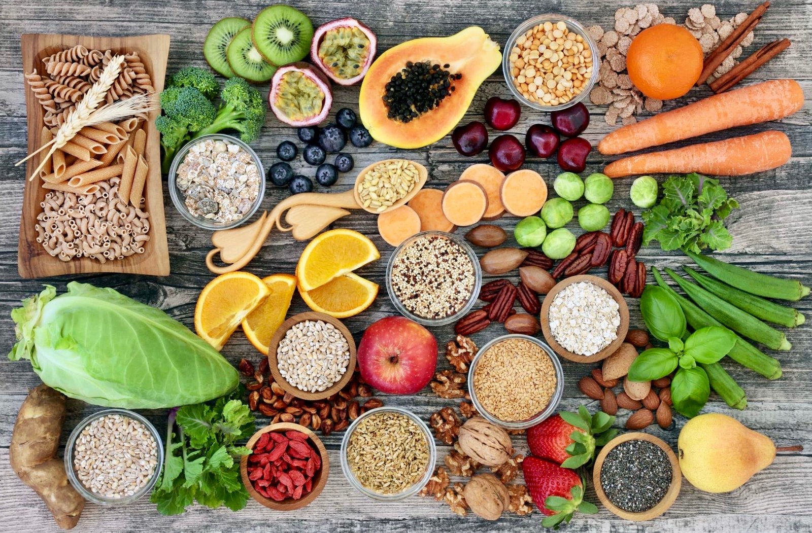 An assortment of real, whole foods including fruits, vegetables, grains, nuts, and herbs on a wooden surface, representing a holistic approach to nutrition.