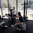 Gym Scene with a Trainer in a White Shirt Guiding a Client Performing Leg-extension on a Seated Machine, Near Large Glass Windows and a Perforated Metal Wall in the Background.