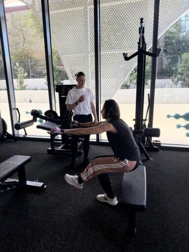 Gym Scene with a Trainer in a White Shirt Guiding a Client Performing Leg-extension on a Seated Machine, Near Large Glass Windows and a Perforated Metal Wall in the Background.