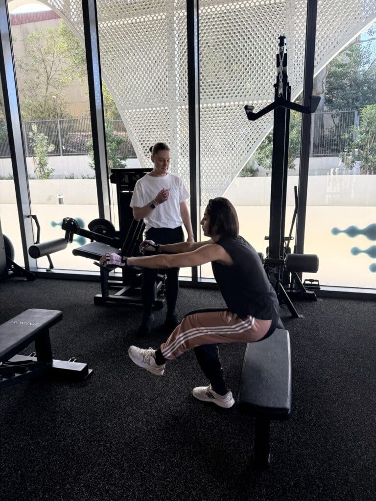 Gym Scene with a Trainer in a White Shirt Guiding a Client Performing Leg-extension on a Seated Machine, Near Large Glass Windows and a Perforated Metal Wall in the Background.